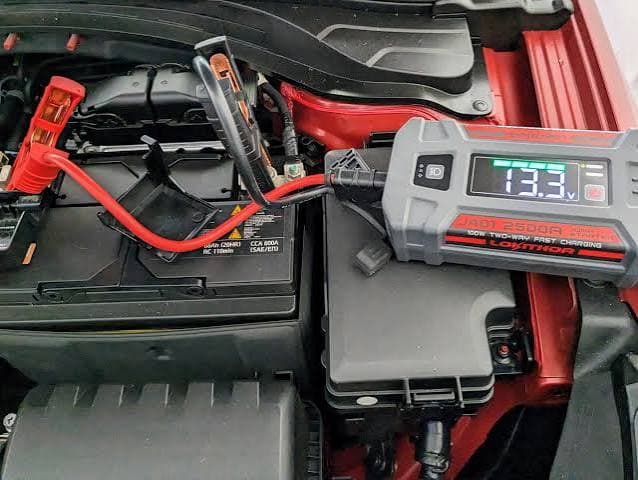 Portable jump starter connected to a car battery, displaying a voltage reading of 13.3V.