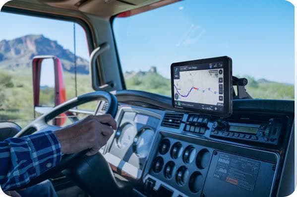 Truck driver's hand on steering wheel with GPS navigation device and scenic desert mountain view.