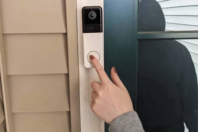 Hand pressing a video doorbell while a hooded figure is visible through the glass door.