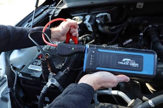 Hands connecting a portable jump starter with red and black clamps to a car battery.